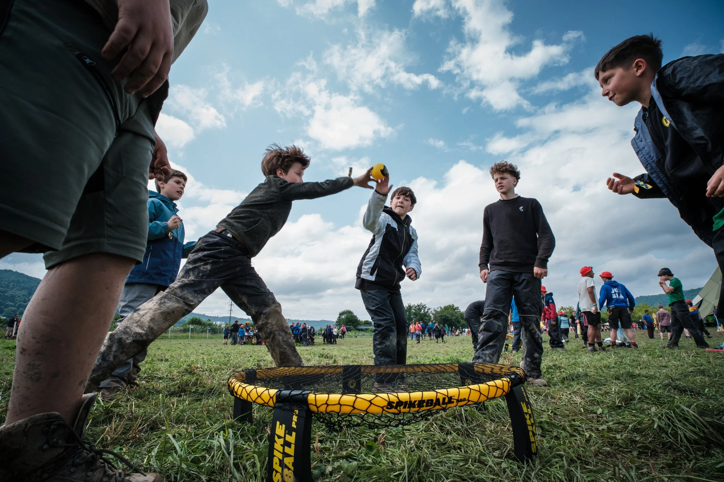 Jugendliche spielen Spikeball im Freien auf einer Wiese, umgeben von anderen Menschen bei einem Event, mit hohen Wolken am Himmel.