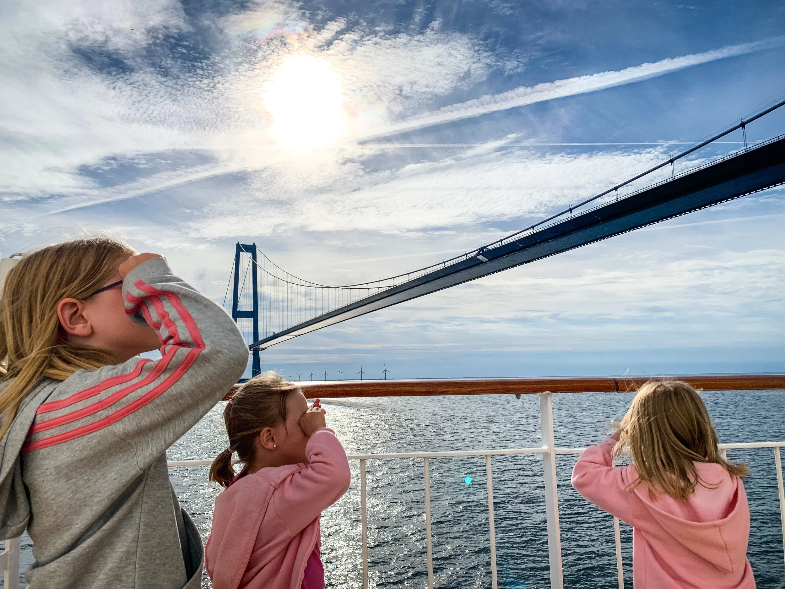 Drei Mädchen auf einem Boot, schauen auf eine Brücke über das Wasser, während die Sonne am Himmel scheint und Wolken und Flugzeugspuren sichtbar sind.