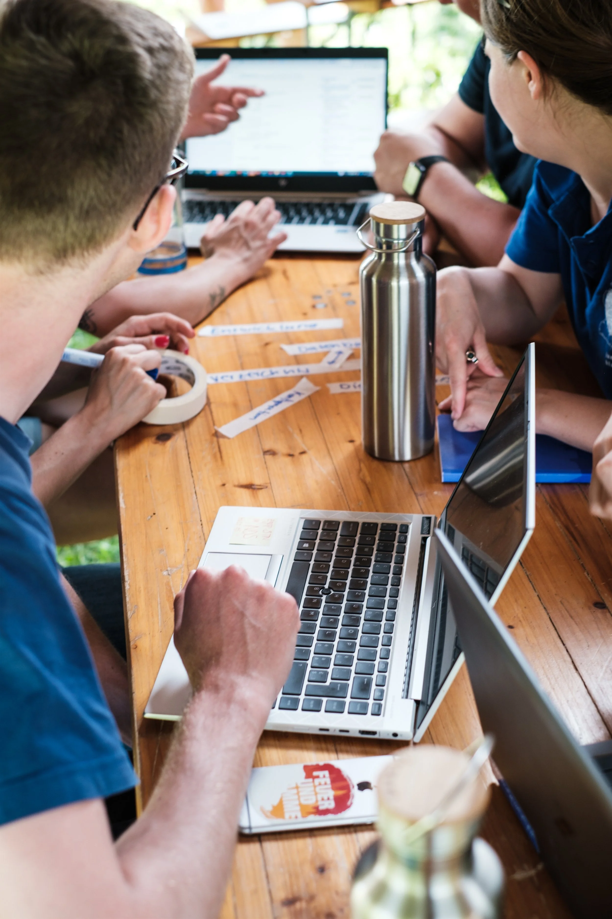 Eine Gruppe von Menschen arbeitet an einem Tisch mit Laptops, Notizen und Trinkflaschen, vermutlich bei einem Treffen oder Workshop im Innenbereich.