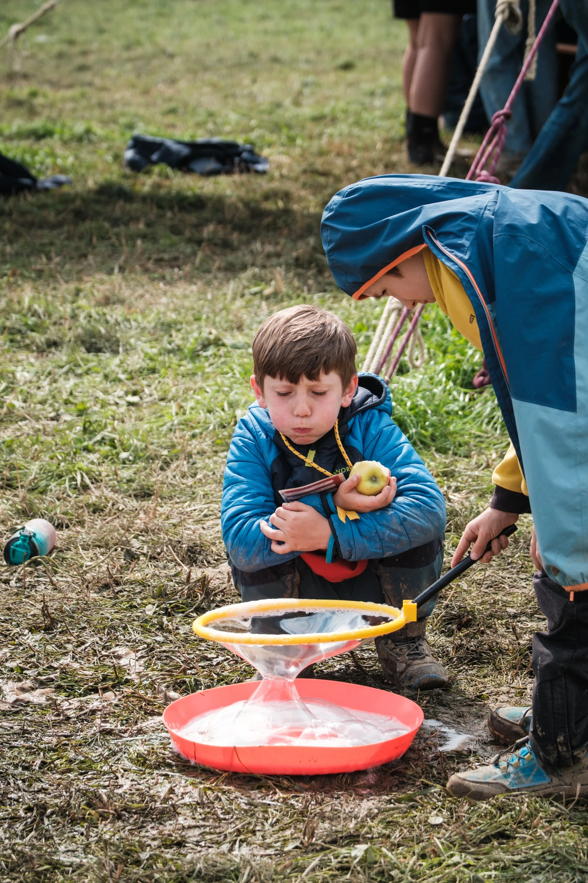 Ein Junge hält einen Apfel und sitzt im Gras während eines Outdoor-Events. Ein anderer Junge benutzt einen Froschfänger, um an einem Seifenblasen-Stand Wasserblasen zu machen. Es sind mehrere Personen im Hintergrund, die an einem Seil ziehen.