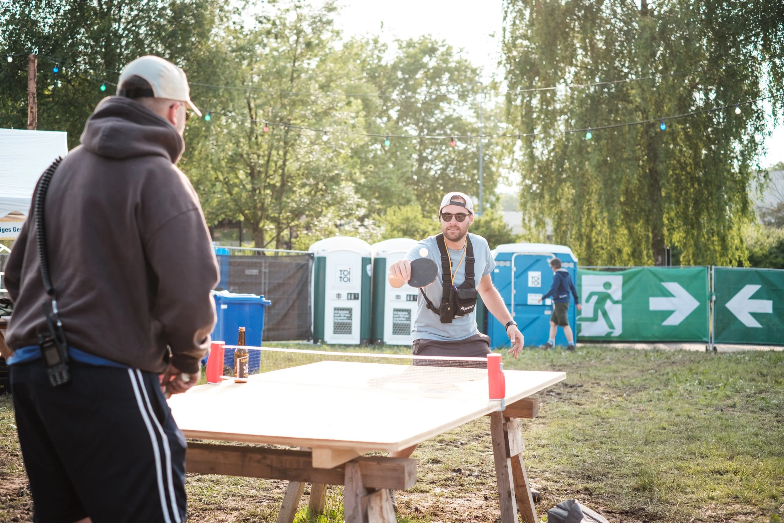 Zwei Männer spielen Tischtennis im Freien, einer schlägt den Ball, der andere beobachtet. Es ist sonnig, im Hintergrund sind Bäume, Zelte und Personen sichtbar.