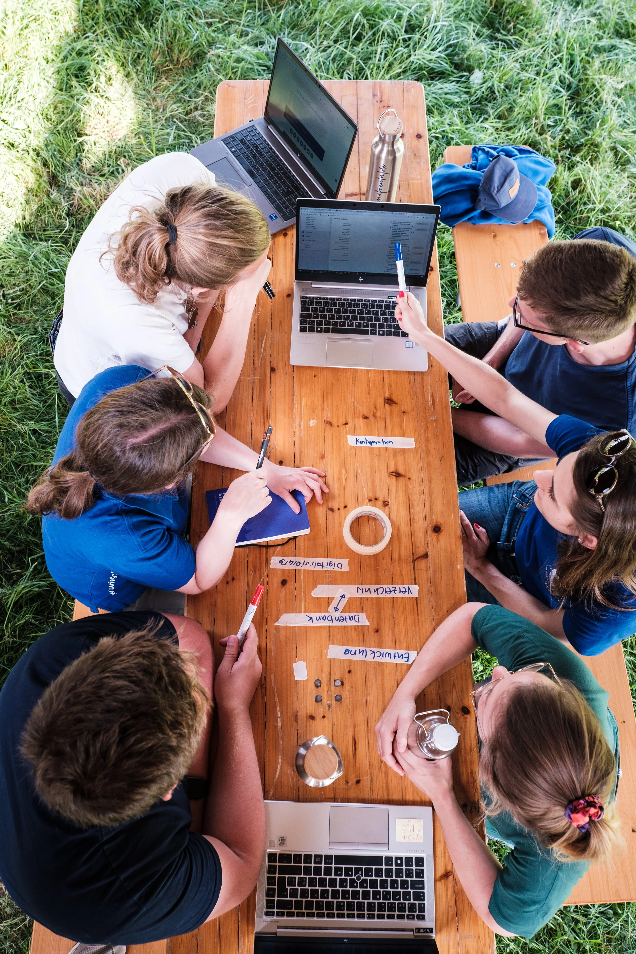 Gruppen von sechs Personen arbeiten zusammen an einem Holztisch im Freien mit Laptops, Notizbüchern, Markern und kleinen Gegenständen, vermutlich bei einem Workshop oder einer Besprechung.