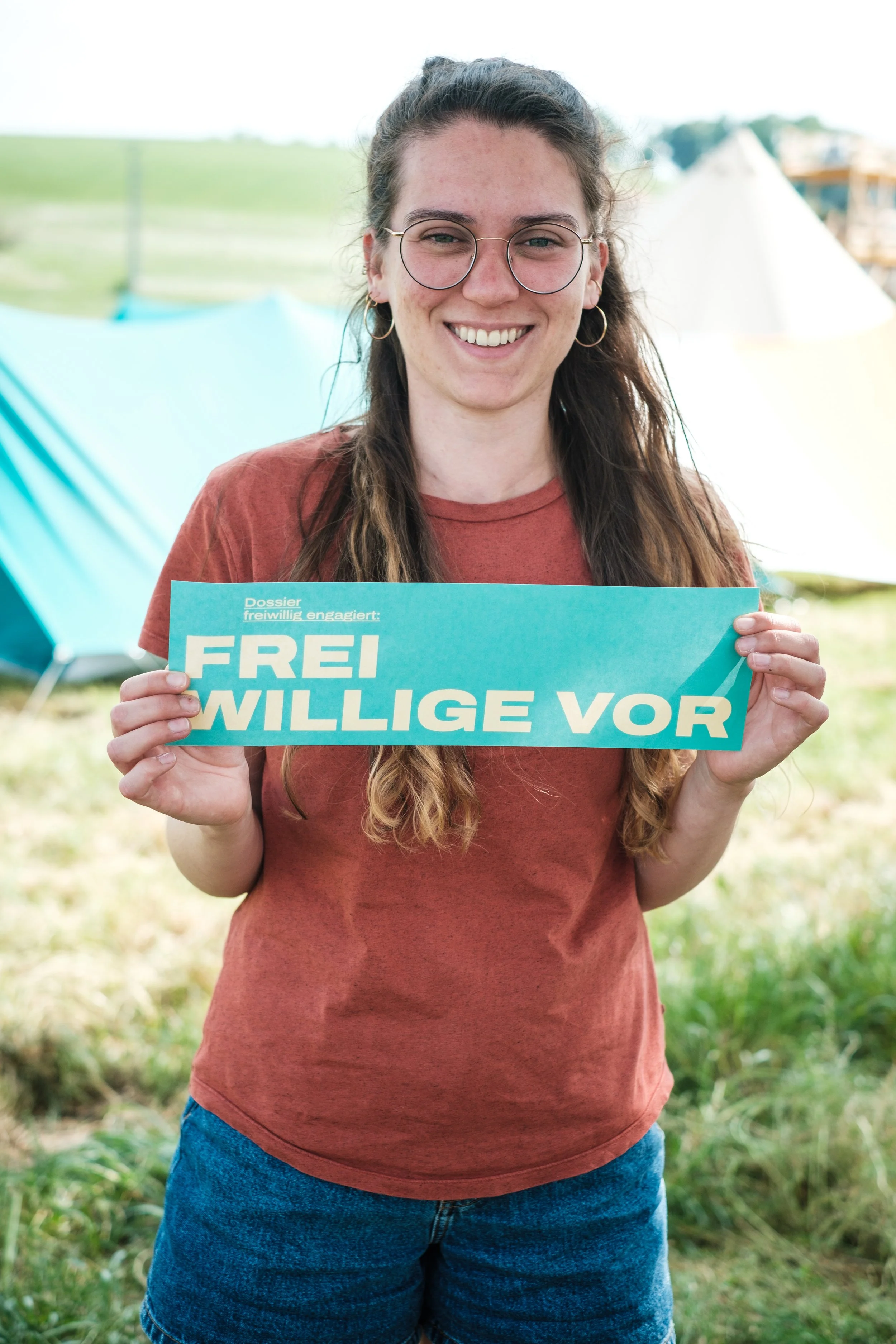 Eine junge Frau hält ein Schild mit der Aufschrift 'FREIWILLIGE VOR'. Sie steht im Freien vor Zelten bei einem Campingplatz.