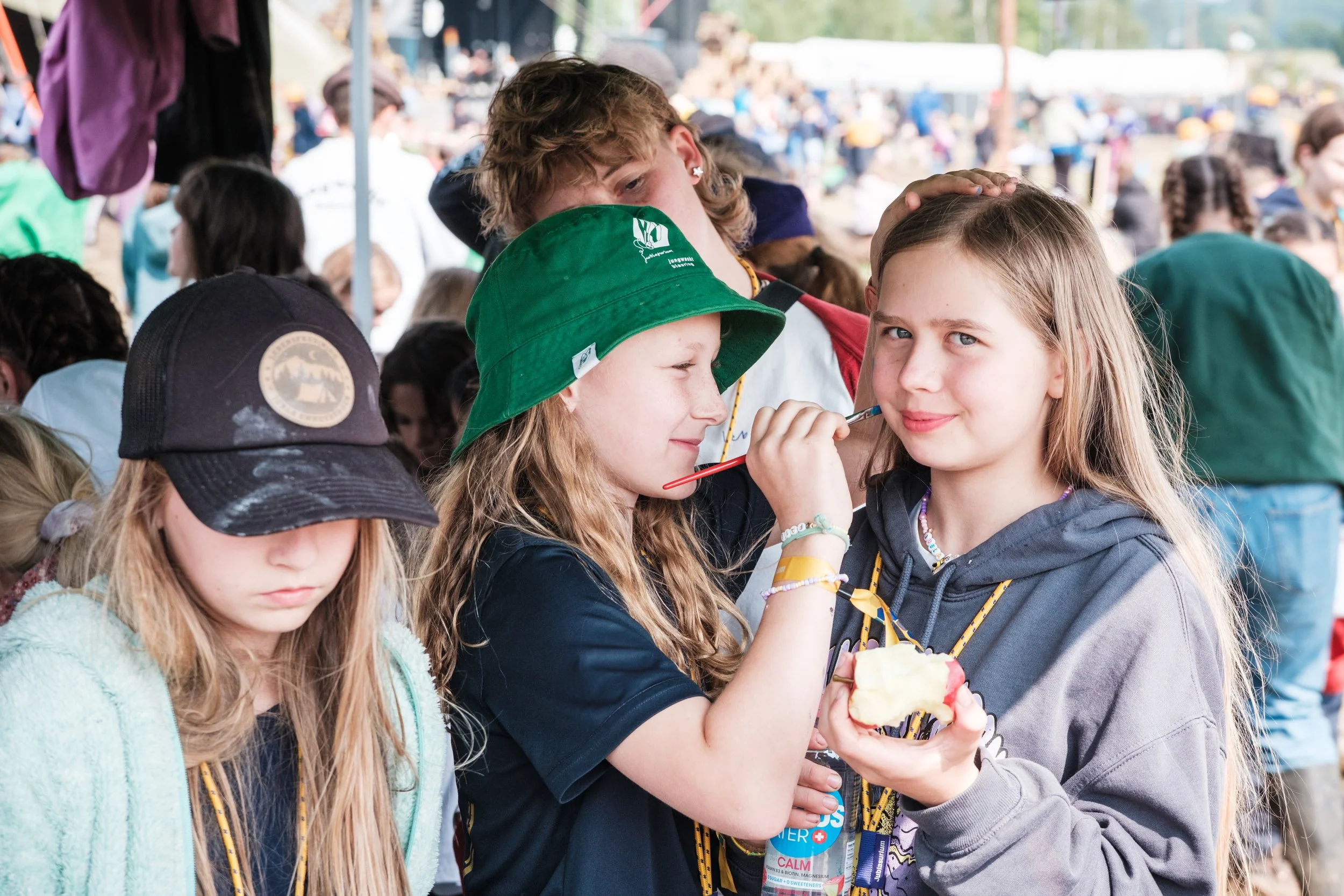 Drei Mädchen auf einem Fest, eines isst Eis, das andere lächelt in die Kamera, alle tragen Lanyards und befinden sich in einer Menschenmenge im Freien.