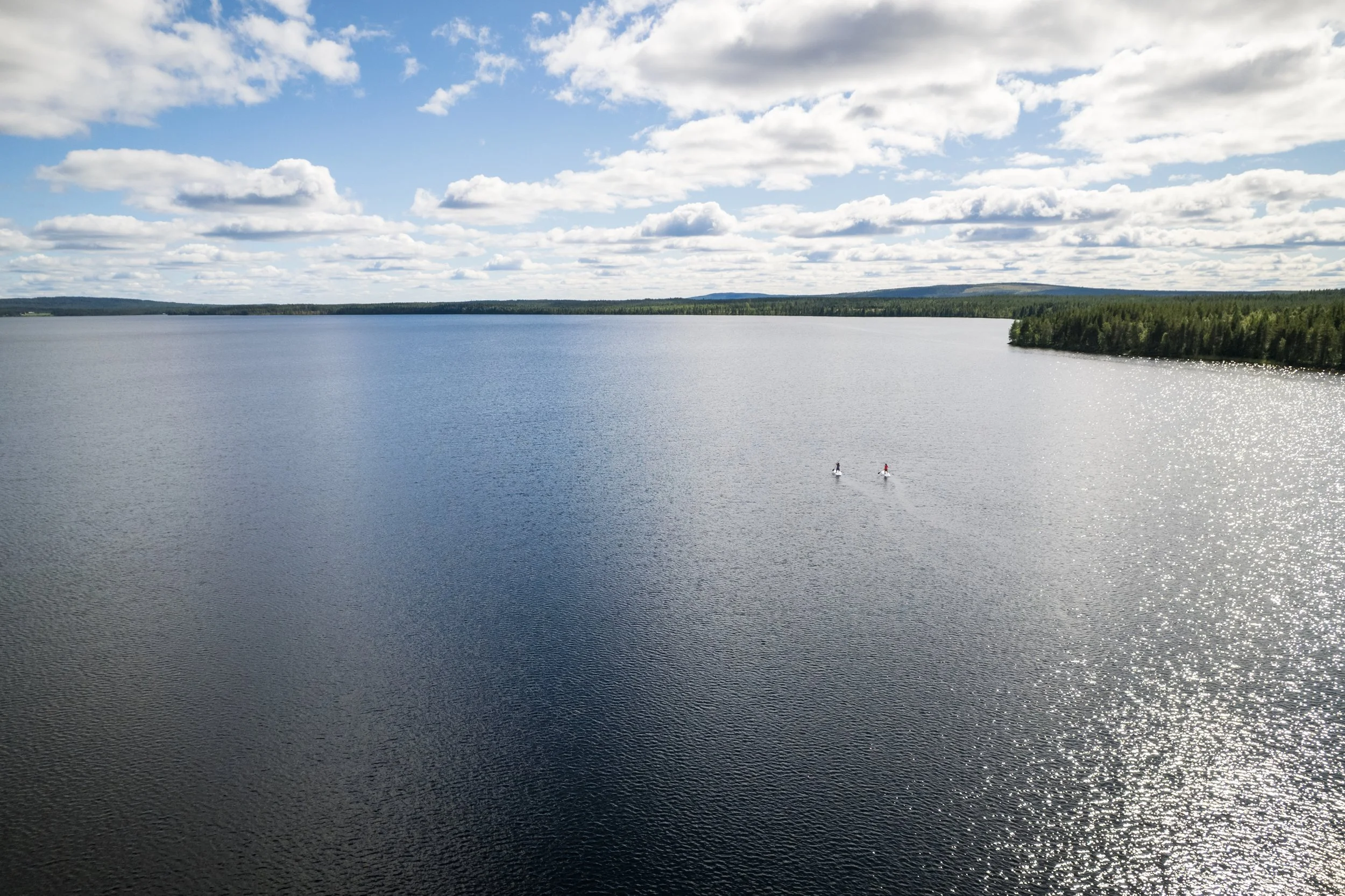 Blick auf einen großen See mit zwei Personen auf Stand-up-Paddeln, umgeben von einem Wald und bei bewölktem Himmel mit Sonne, die auf das Wasser reflektiert.