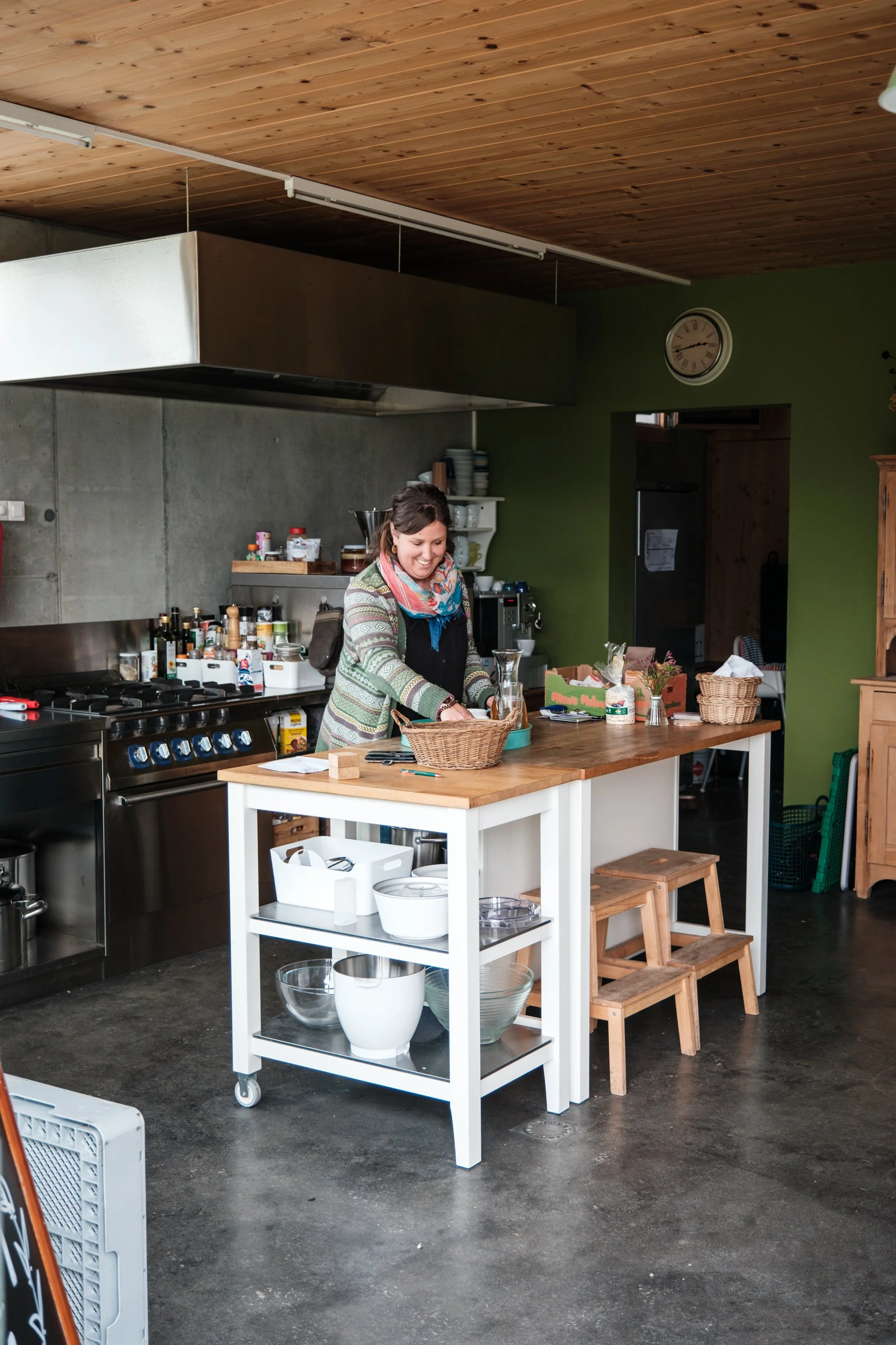Eine Frau in einer Küche beim Kochen oder Vorbereiten, umgeben von Küchenutensilien und Lebensmitteln, mit einem grünen Wandhintergrund und Holzakzentdecken.