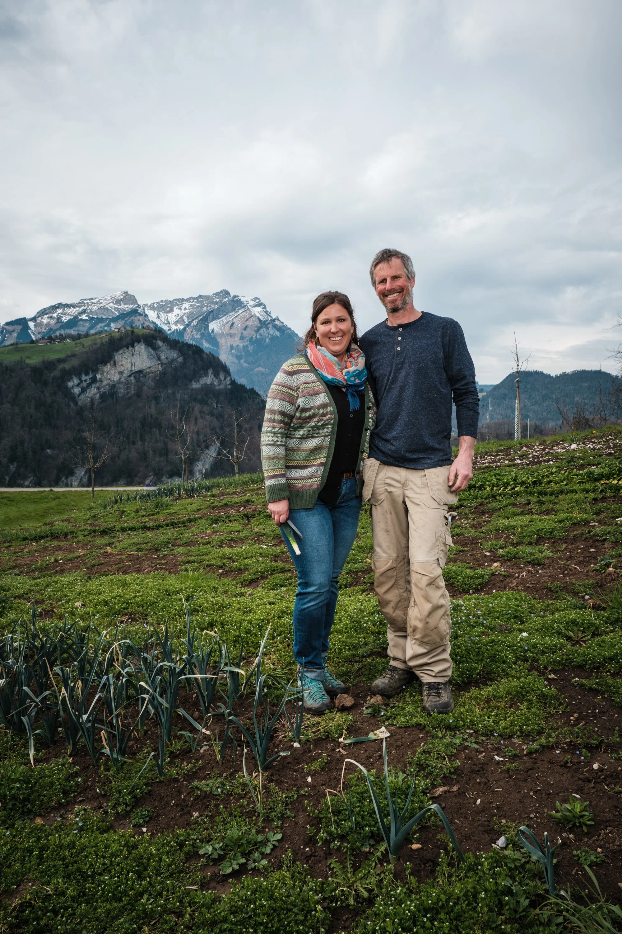 Ein Mann und eine Frau stehen im Feld mit einem Bergpanorama im Hintergrund, lächelnd.
