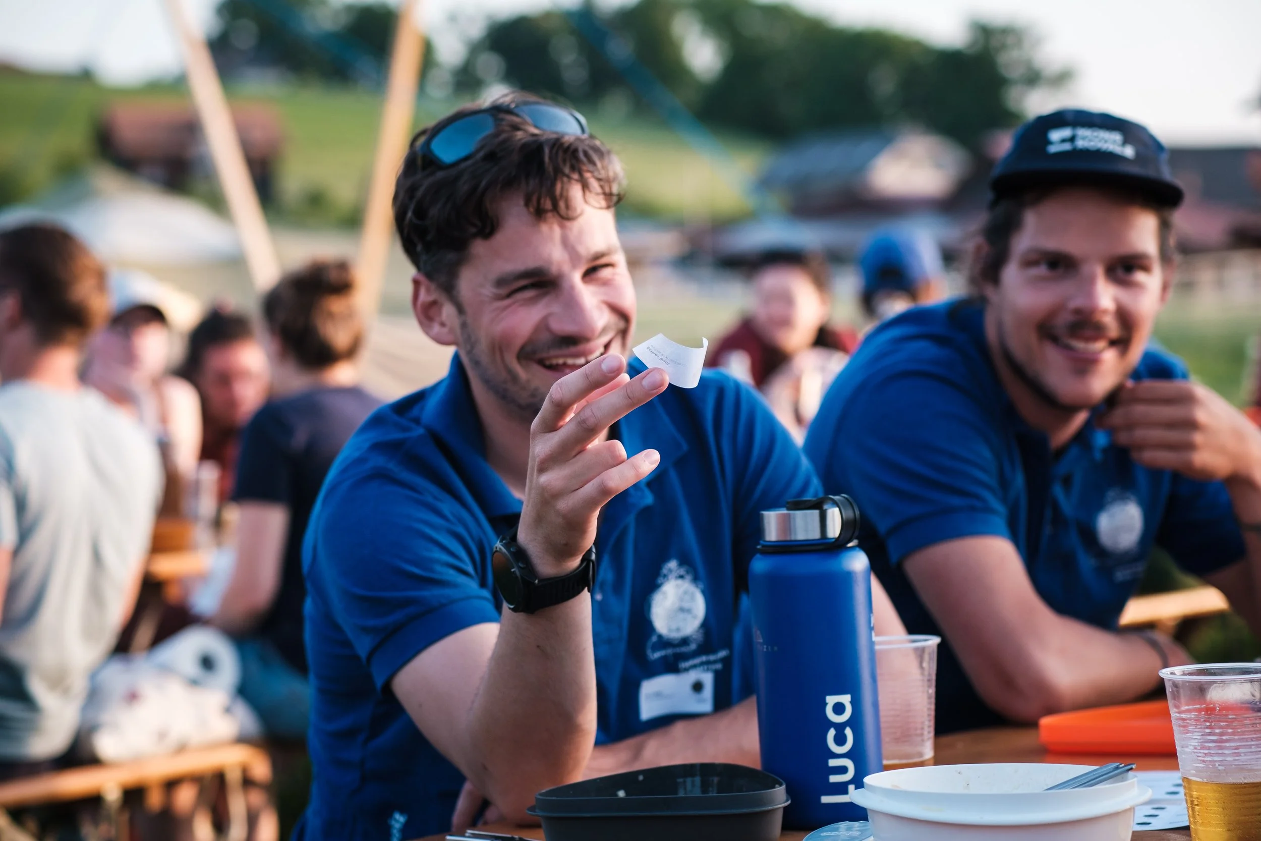 Zwei lachende junge Männer in blauen T-Shirts sitzen an einem Tisch im Freien und genießen eine Mahlzeit bei einem sozialen Event.