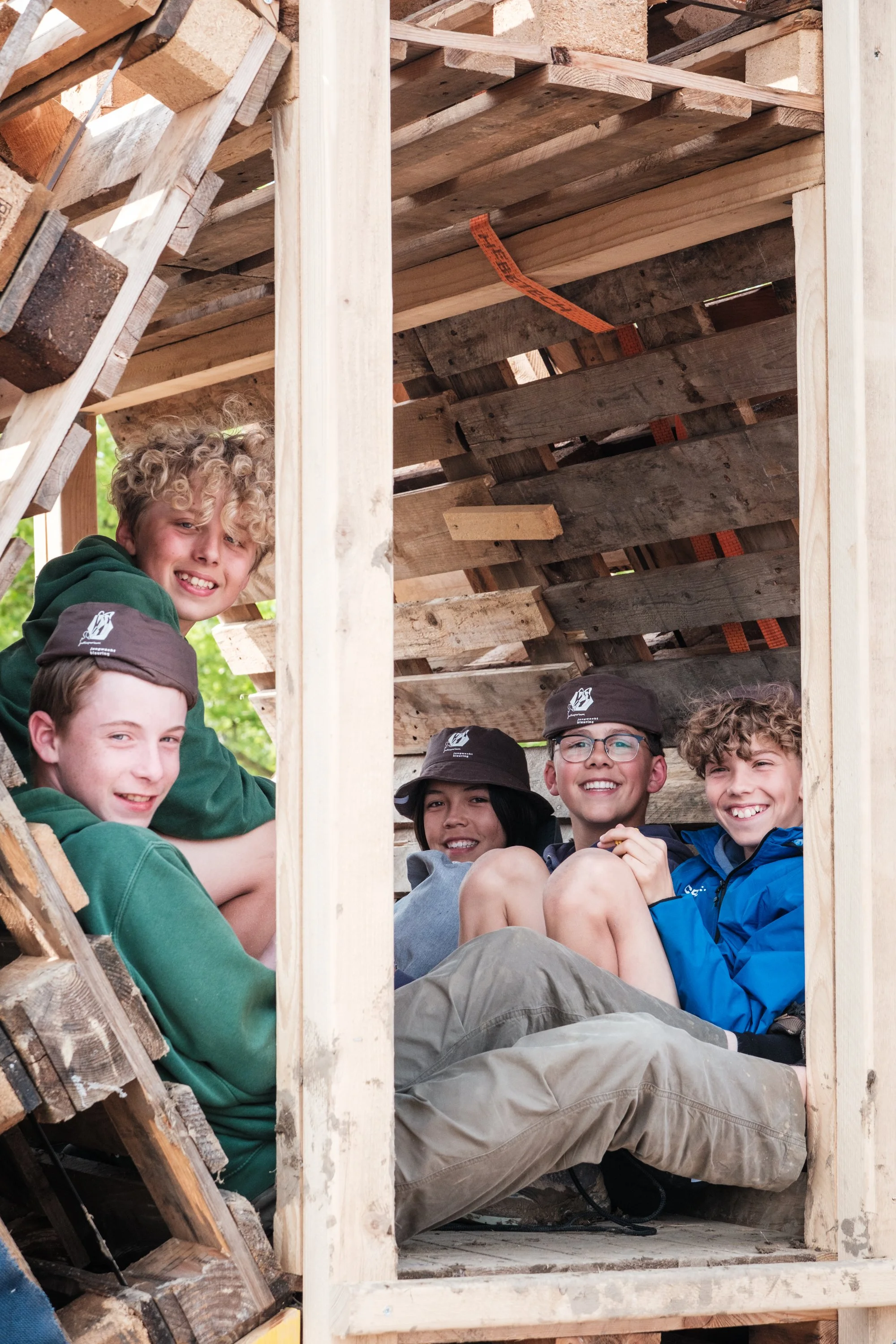 Sechs Kinder sitzen im Inneren eines selbstgebauten Holzhauses aus Paletten, lachen und posieren für die Kamera