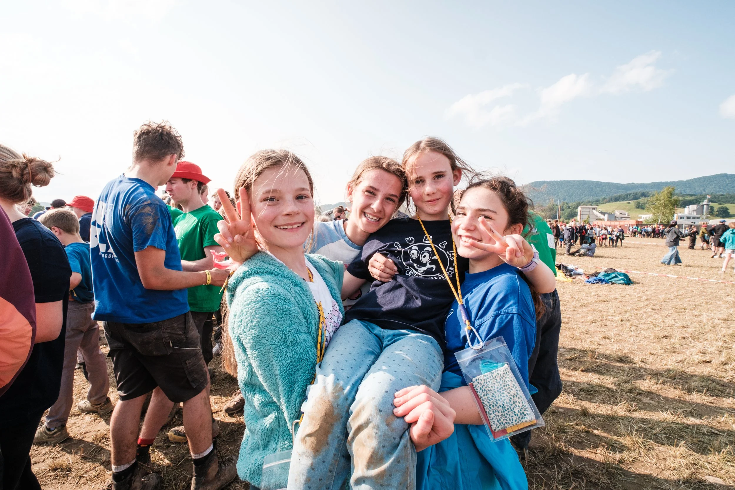 Gruppe lachender Kinder bei einem Outdoor-Event, einige machen Peace-Zeichen, im Hintergrund sind weitere Leute und ein spätsommerlicher Himmel.