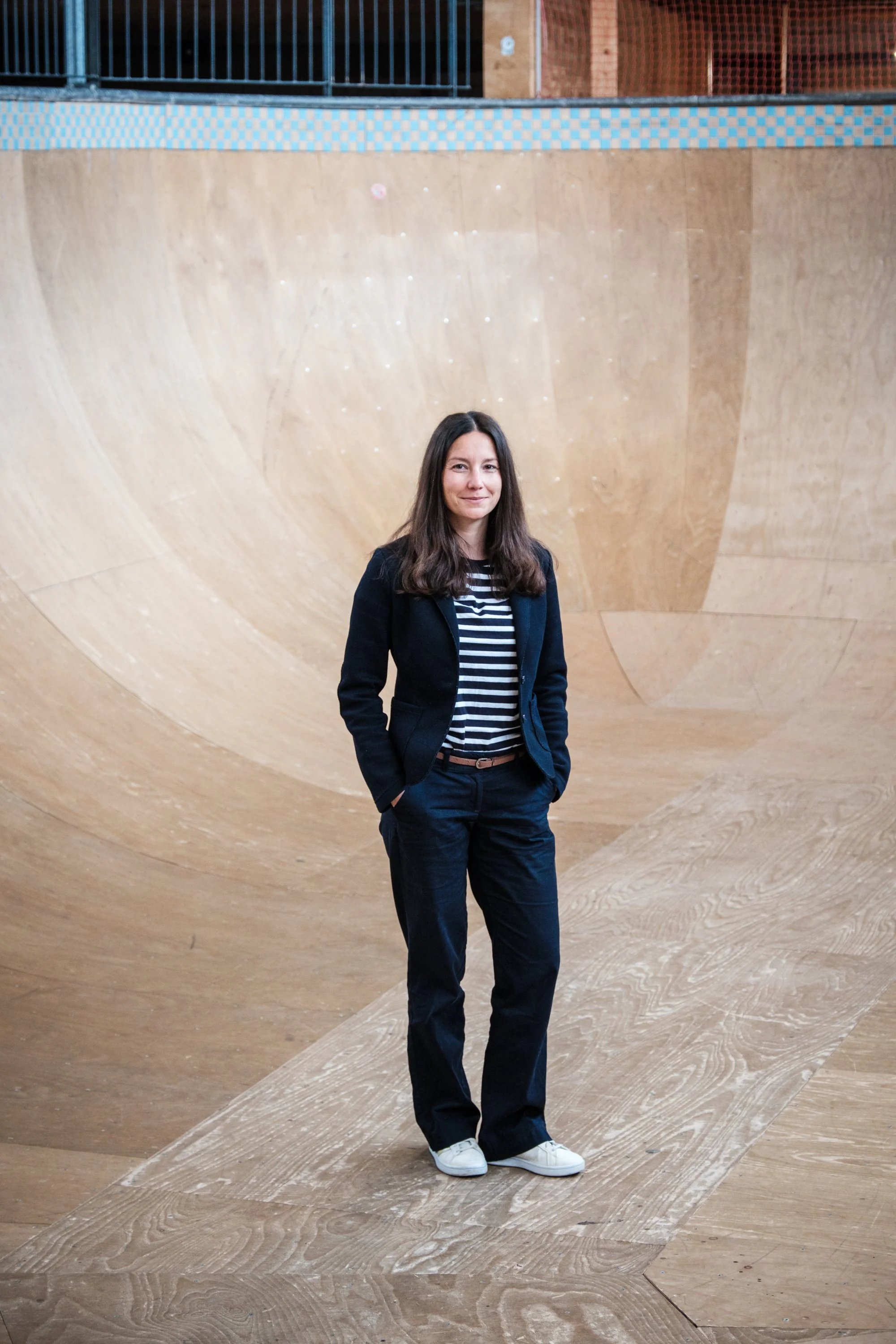 Eine Frau steht in einem Skatepark mit einer großen Holzrampe im Hintergrund, trägt dunkle Kleidung und hat braune Haare.
