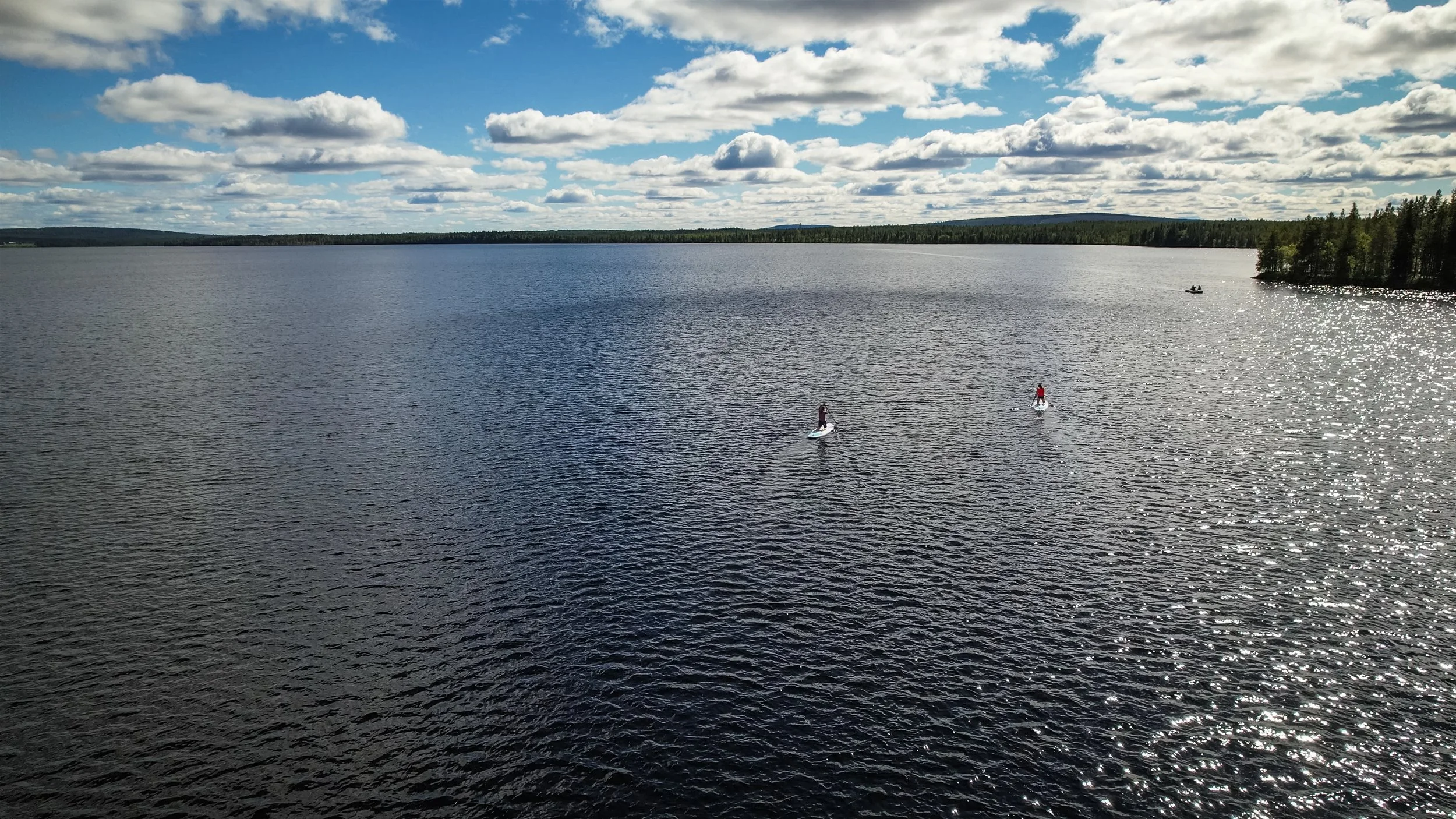 Blick auf einen großen See mit zwei Personen, die auf Stand-up-Paddle-Boards fahren, und einem kleinen Boot im Hintergrund; bewölkter Himmel bei Tageslicht.
