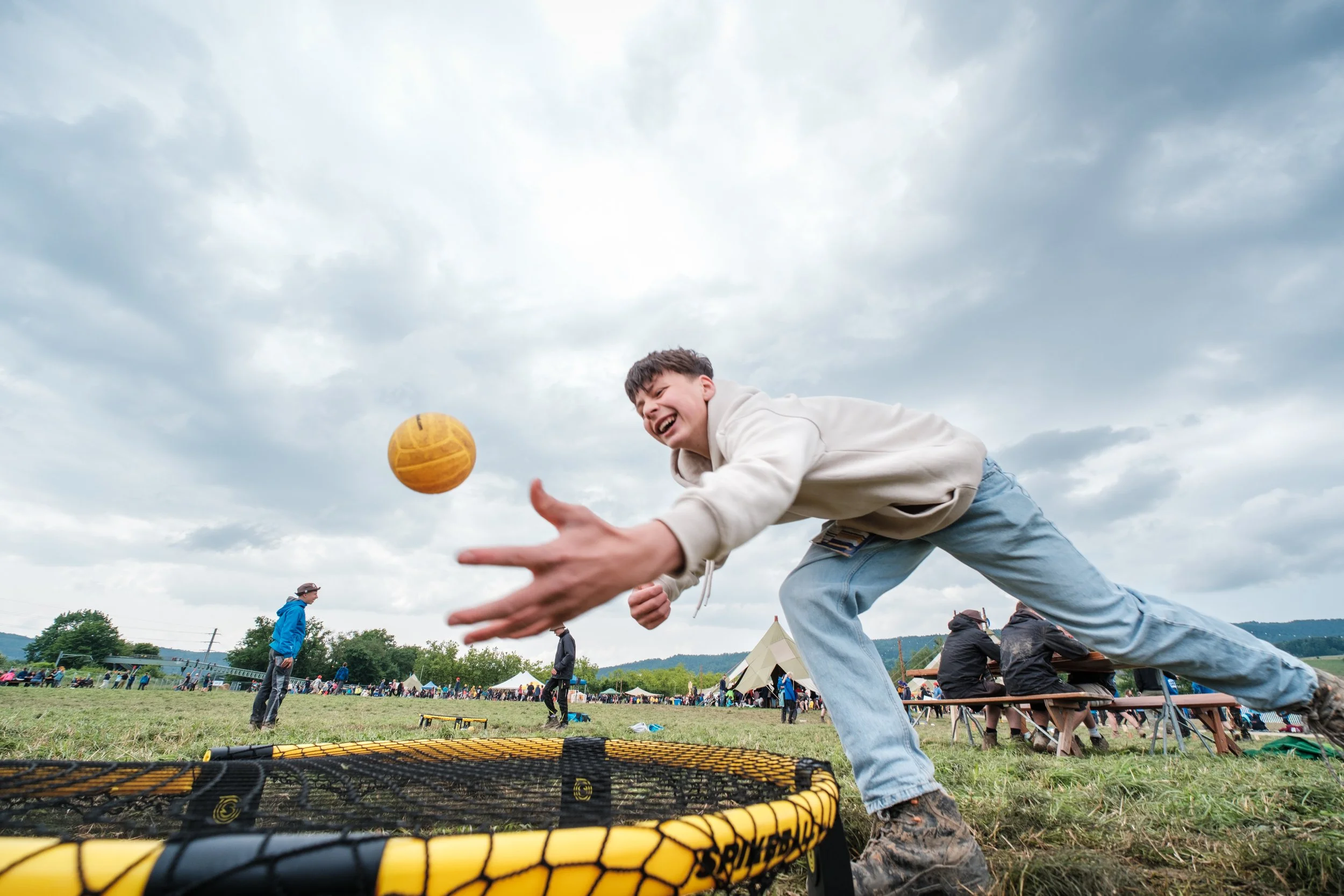 Junge beim Basketballwerfen in einem Freiluftpark, mit Zelten im Hintergrund und Wolken am Himmel.