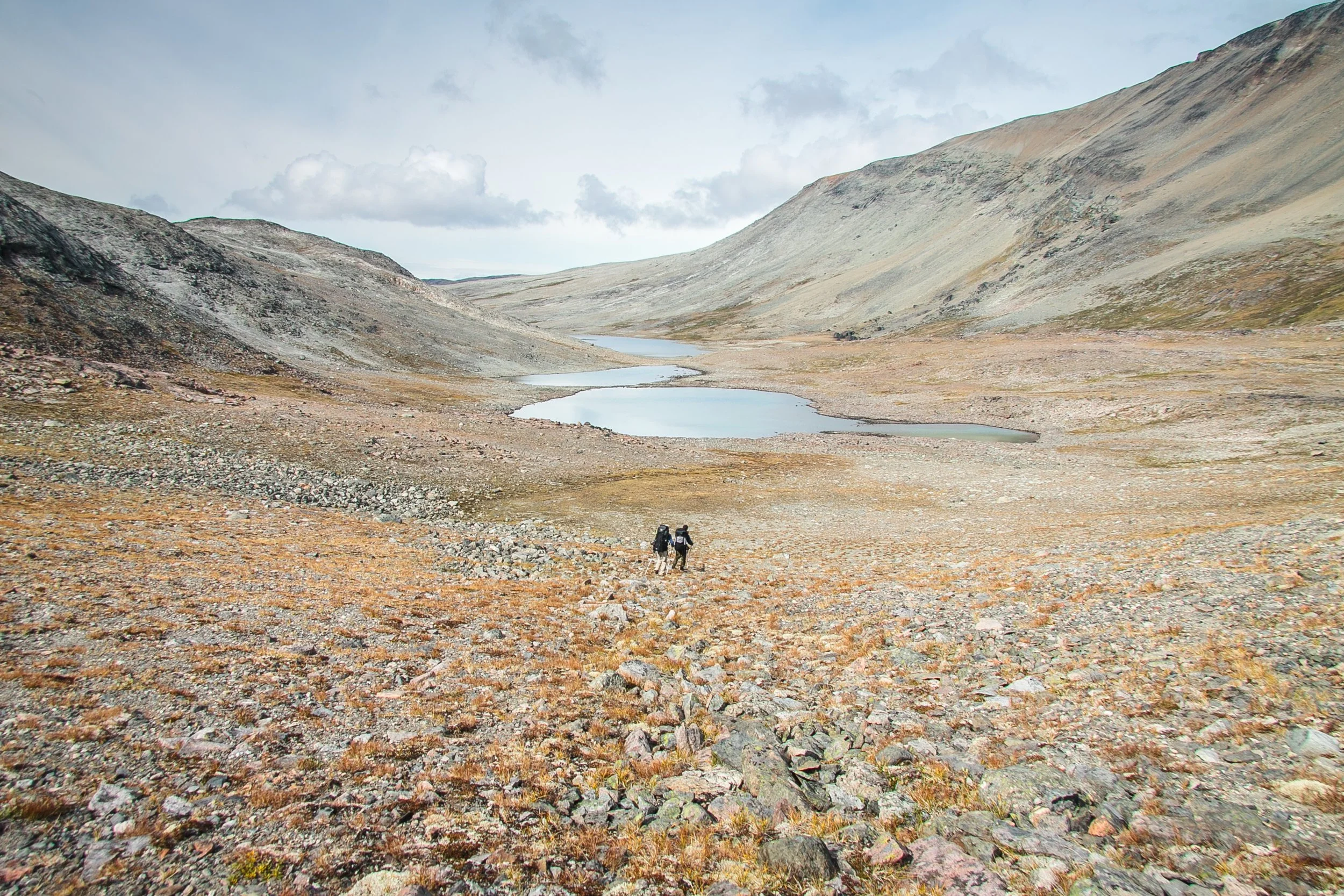 Zwei Wanderer inmitten einer kargen, bergigen Landschaft mit kleinen Seen und steinigem Terrain.