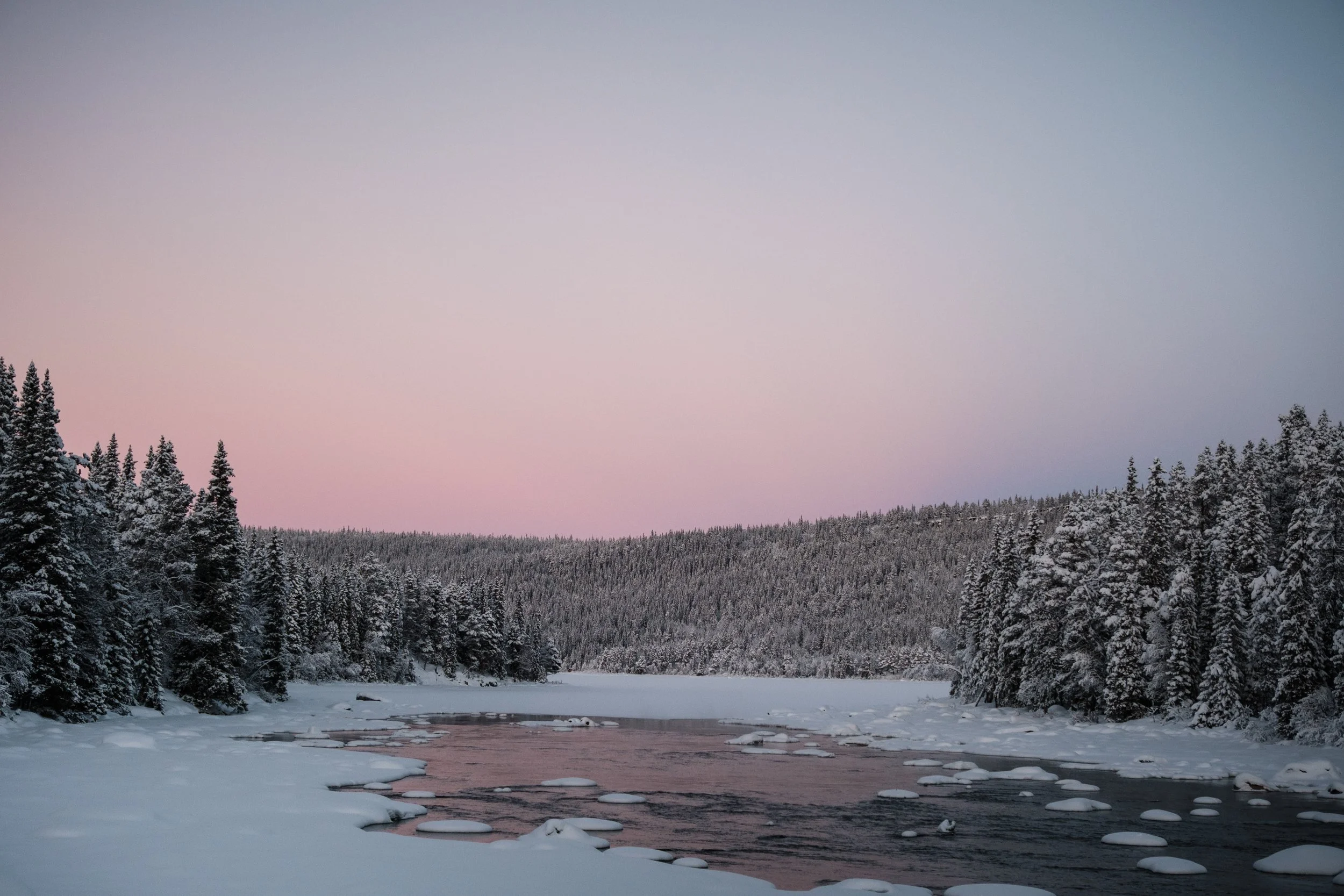 Schneebedeuer Wald am Fluss bei Sonnenaufgang im Winter.