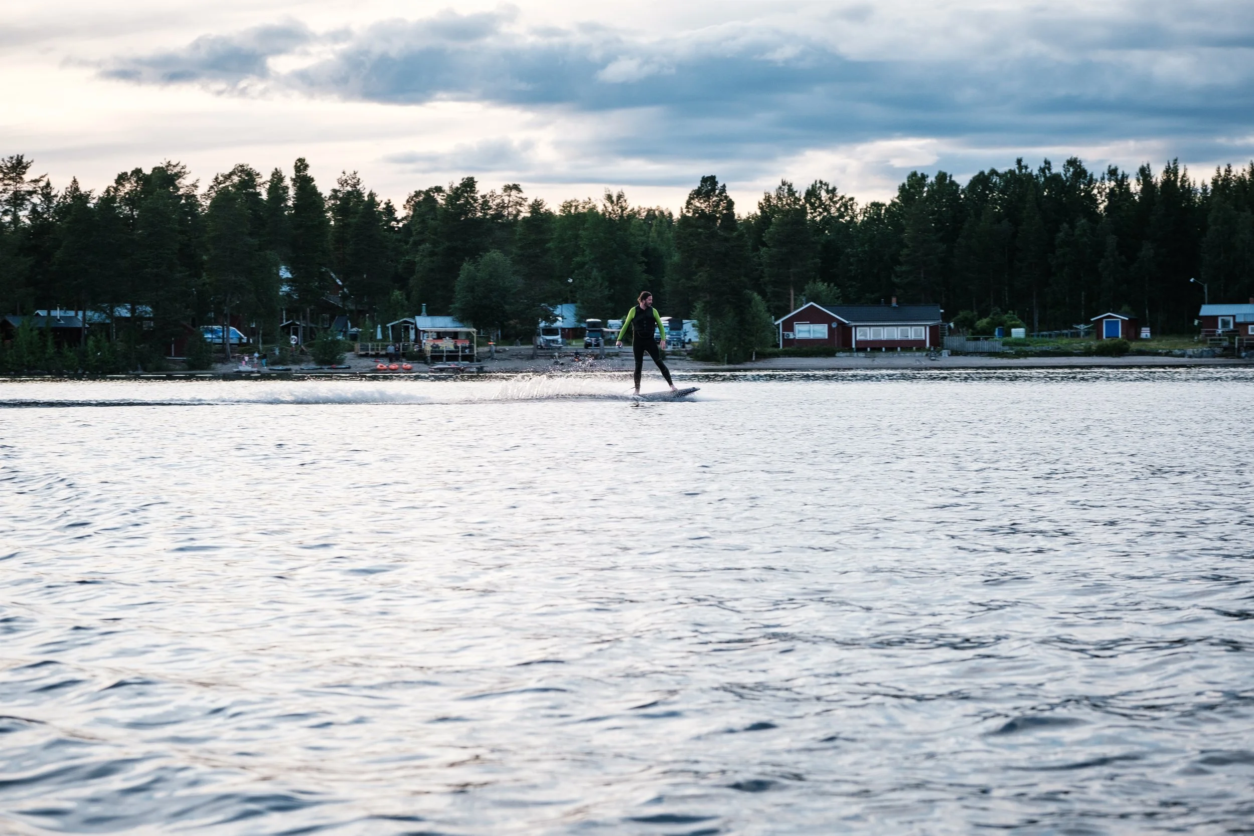 Mensch beim Wasserskifahren auf einem See mit Häusern und Bäumen im Hintergrund, bewölkter Himmel.