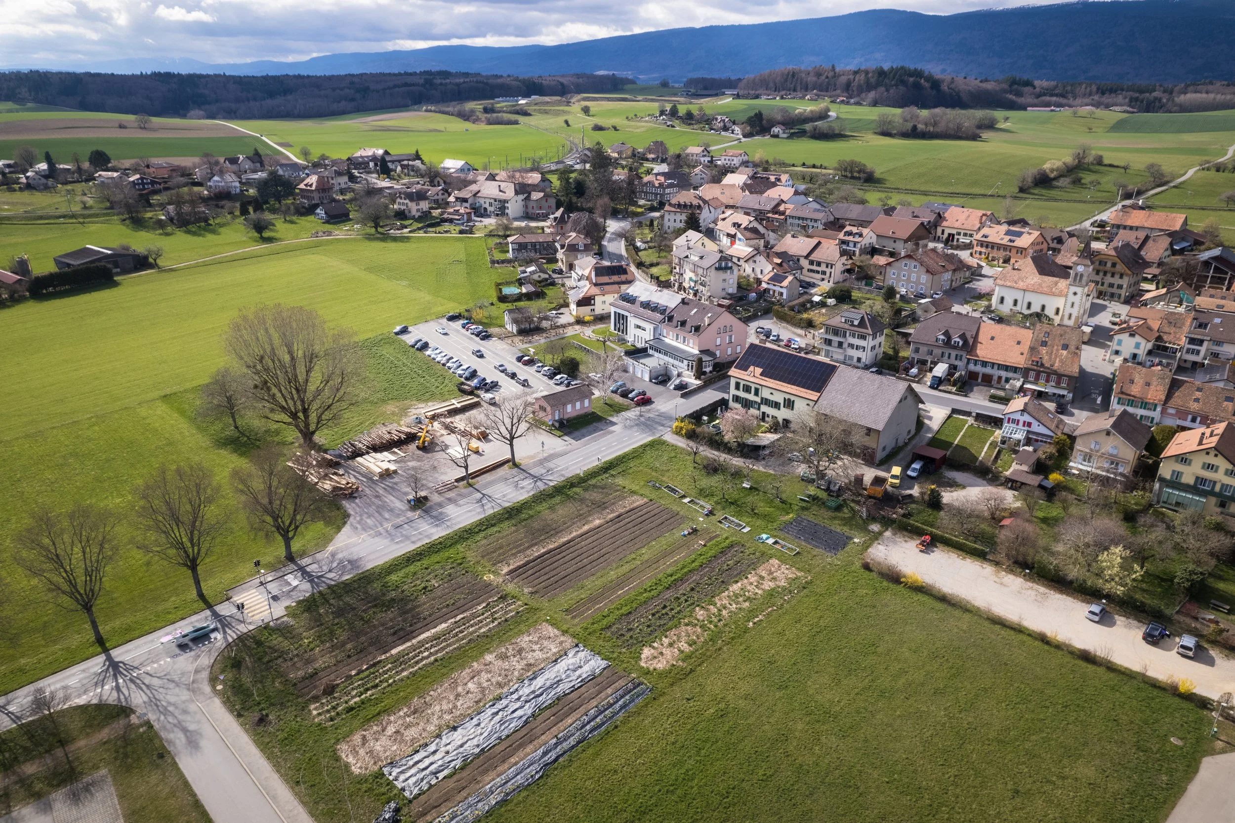Luftaufnahme eines kleinen Dorfes mit Wohnhäusern, Feldern, einem Parkplatz und landwirtschaftlichen Flächen in einer grünen, hügeligen Landschaft mit Bergen im Hintergrund.