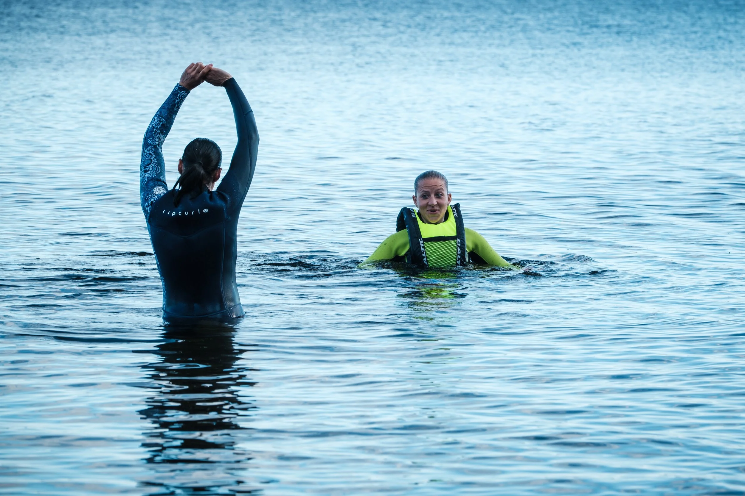 Zwei Frauen im Wasser, eine übt Yoga, die andere trägt eine Schwimmweste und spricht.