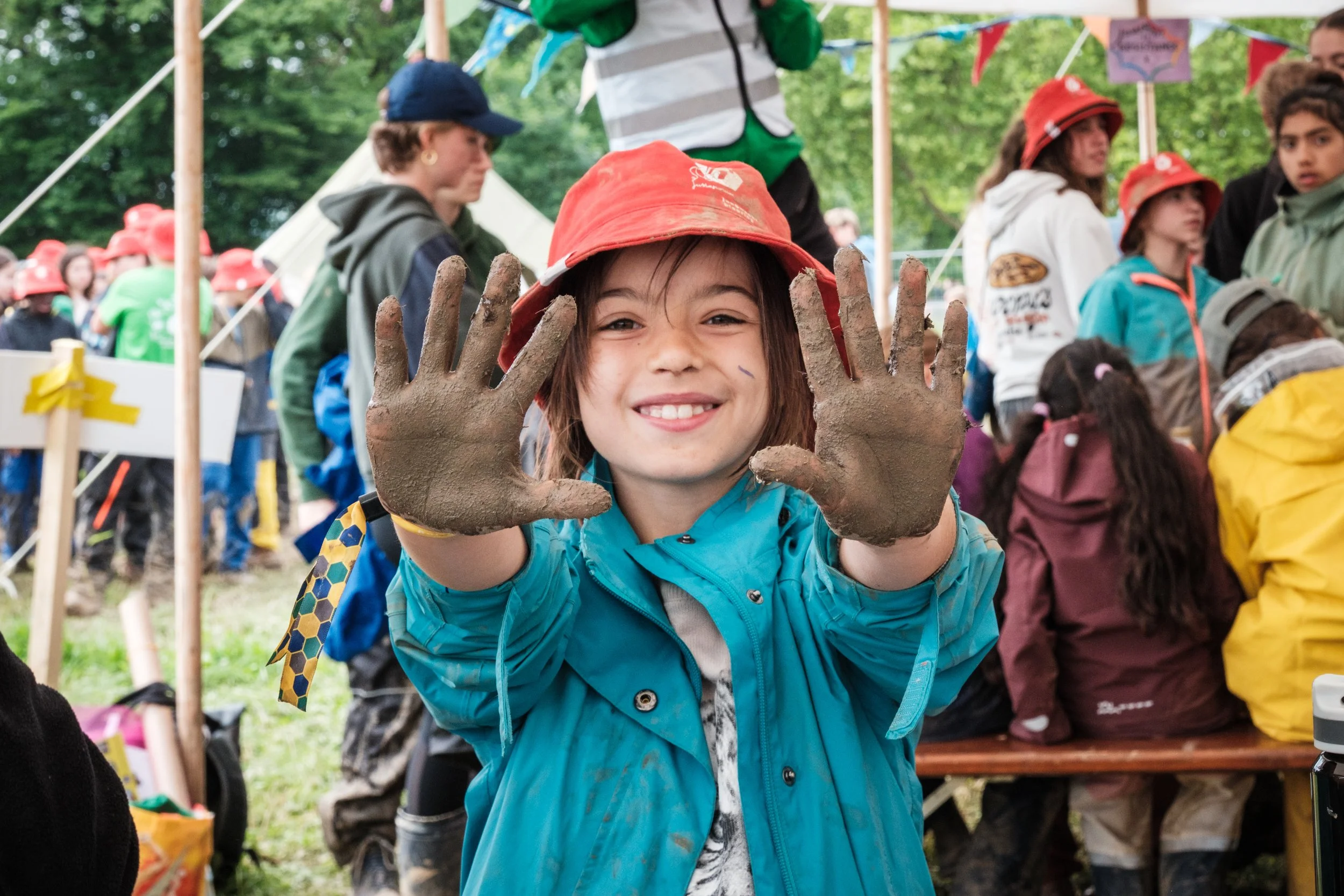 Ein glückliches Kind mit bemalten Händen bei einer Gruppenschulveranstaltung im Freien, umgeben von anderen Kindern und Erwachsenen.