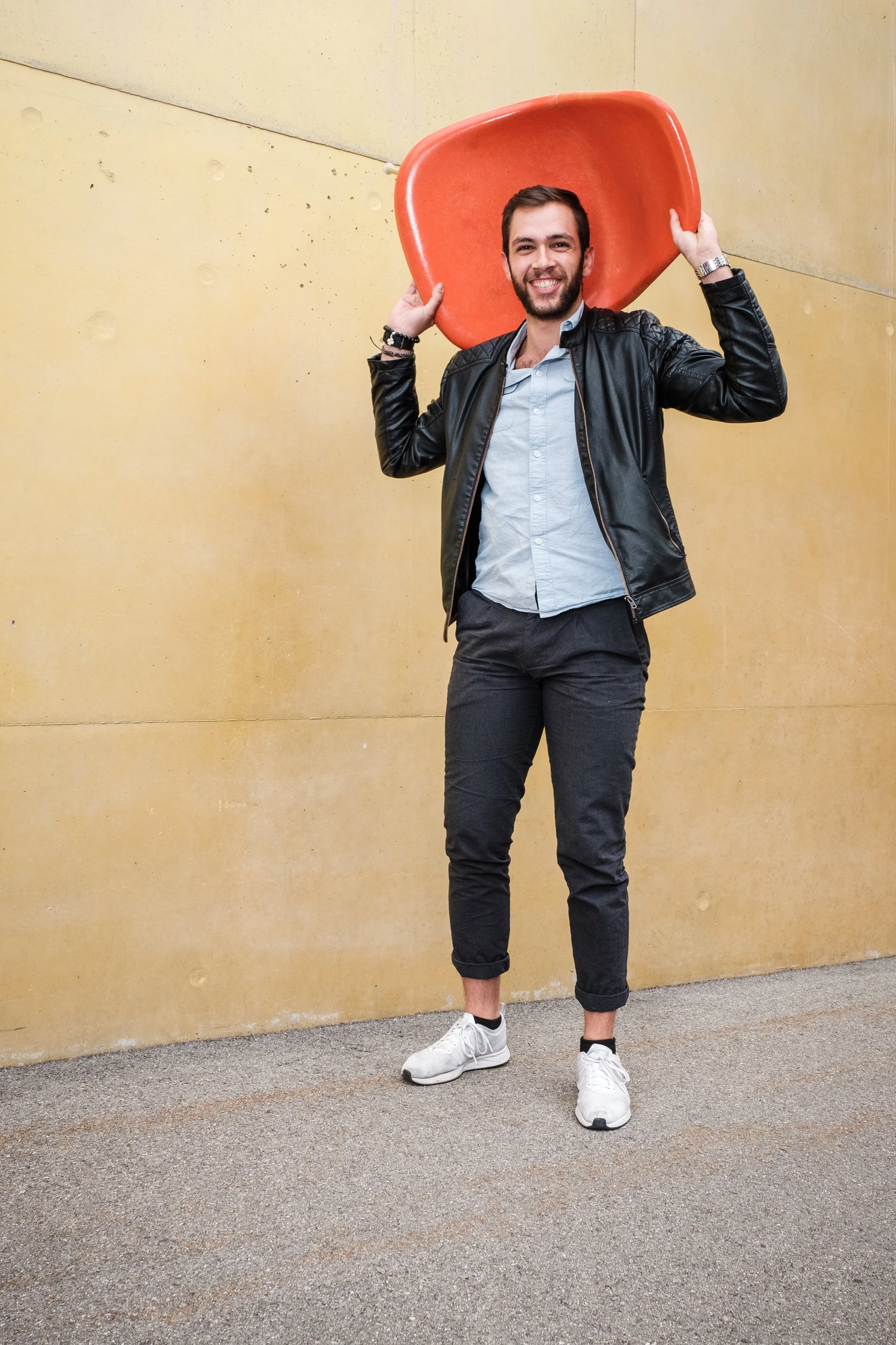 Ein lächelnder junger Mann trägt eine schwarze Lederjacke, eine helle Hemd, schwarze Hosen, weiße Sportschuhe und hält einen roten Stuhl auf den Schultern, steht vor einer gelben Wand.