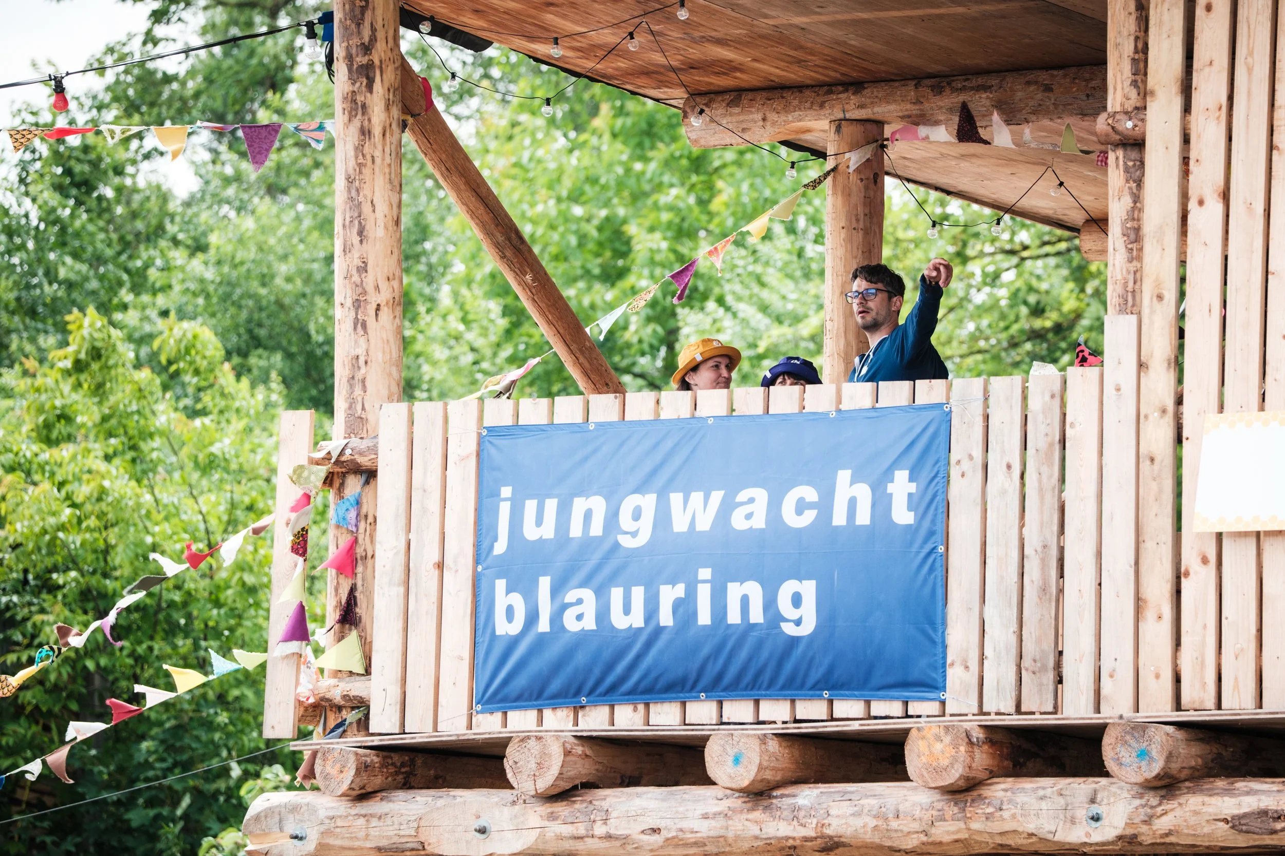 Menschen auf einer Holzplattform, die durch bunte Flaggen und Lichter dekoriert ist, mit einem blauen Banner, auf dem 'jungwacht blauring' steht.