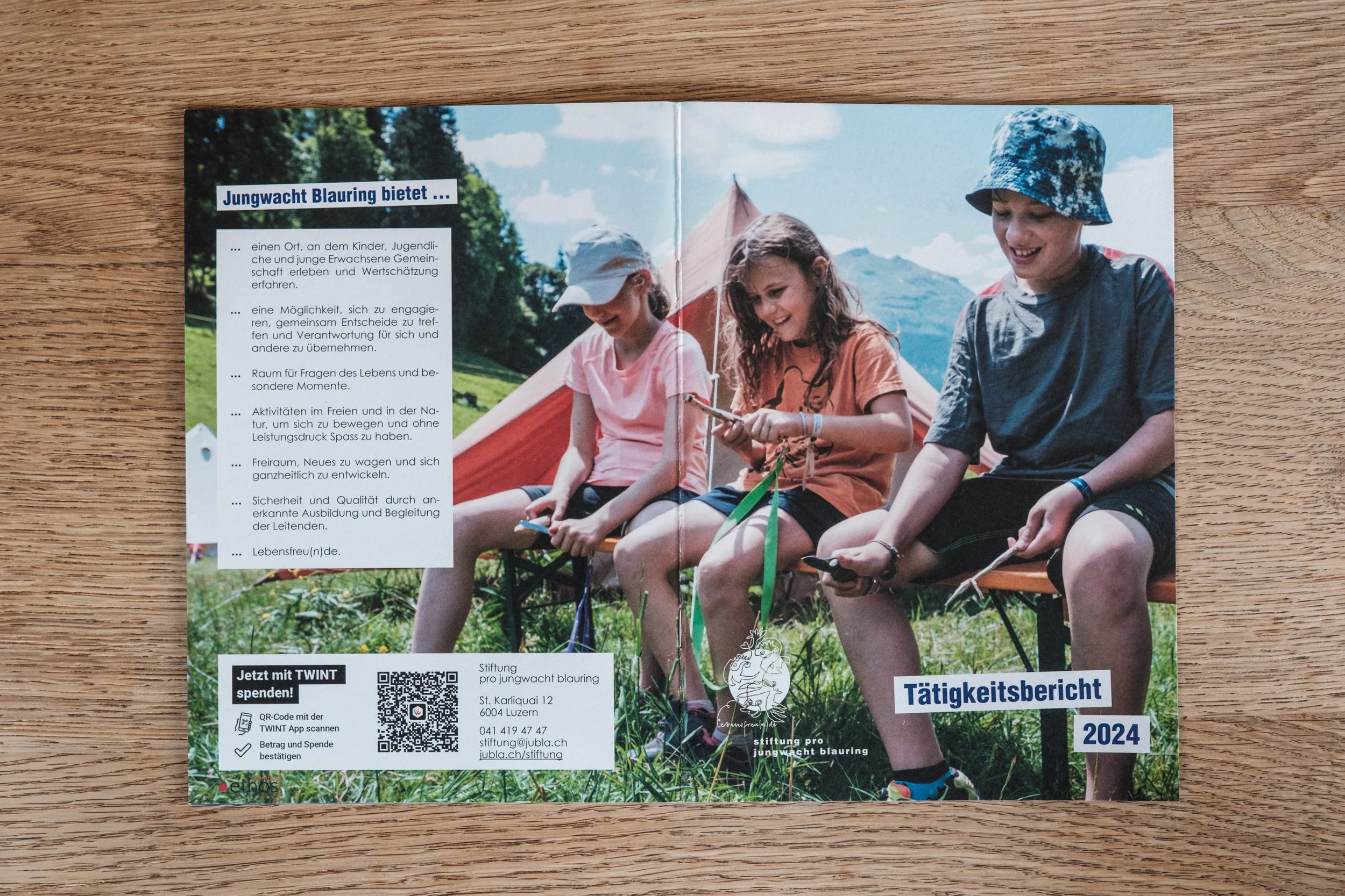 Bild eines Aktivitätenberichts für 2024 der Stiftung pro jungwacht blauring mit drei lachenden Kindern, die im Freien auf einem Stuhl sitzen, vor einer Zelthütte und Bergen im Hintergrund, bei sonnigem Wetter.