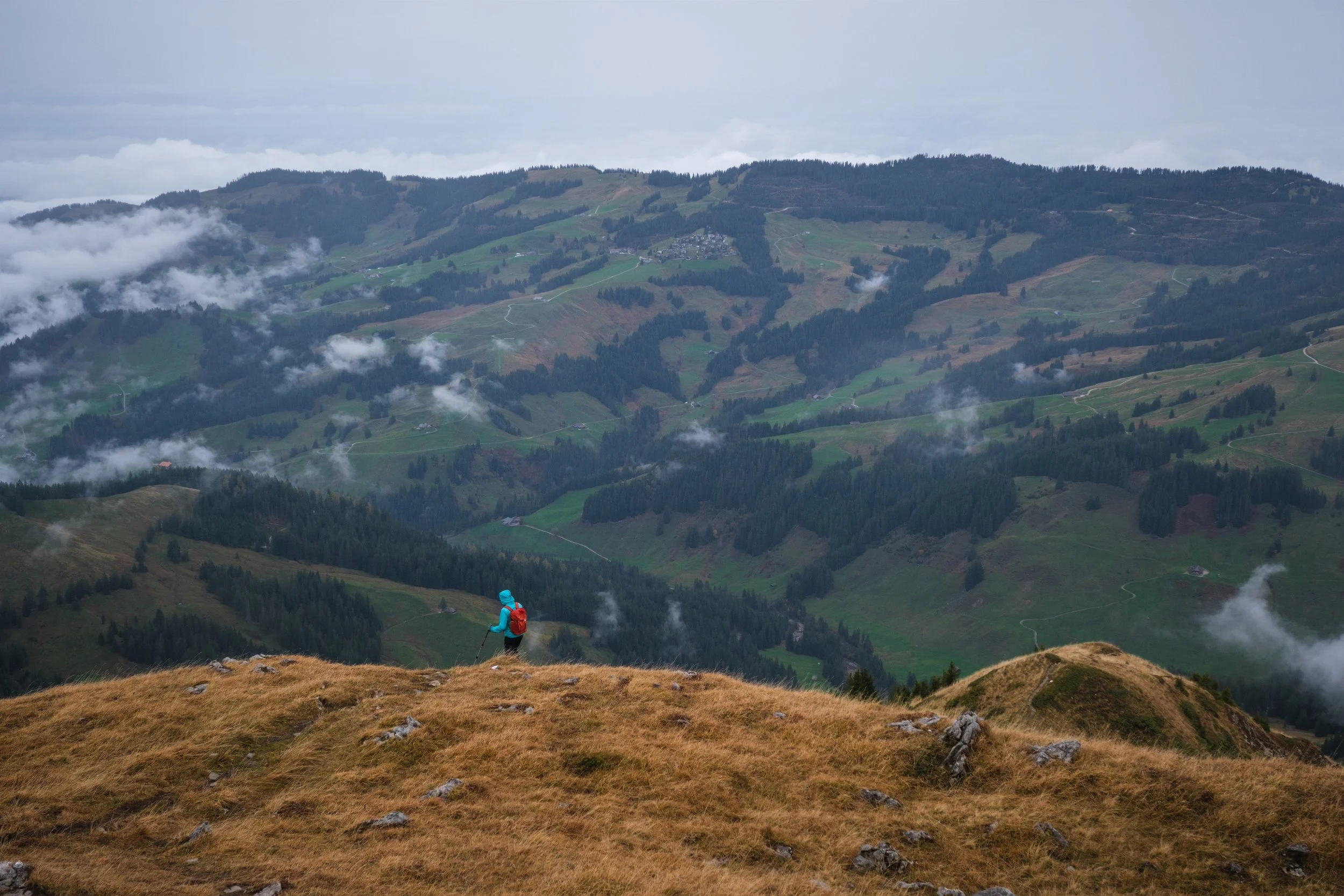 Ein Bergsteiger mit blauer Jacke und rotem Rucksack, wandert auf einem grasbedeckten Gipfel mit Blick auf eine niederschlagsreiche Berglandschaft mit Tälern und Wäldern.