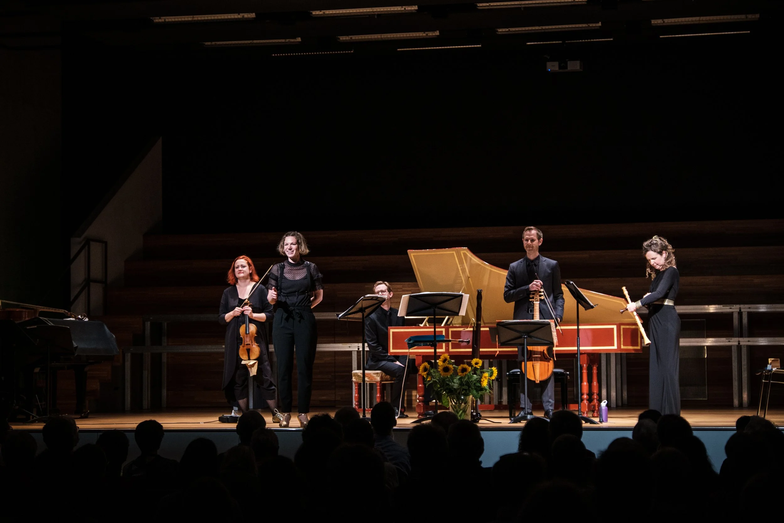 Musiker auf einer Bühne beim Konzert, mit Streichinstrumenten, Klavier, Cello, Flöte und Harfe, Publikum im Vordergrund, dunkler Hintergrund, Sonnenblumen in der Mitte der Bühne.
