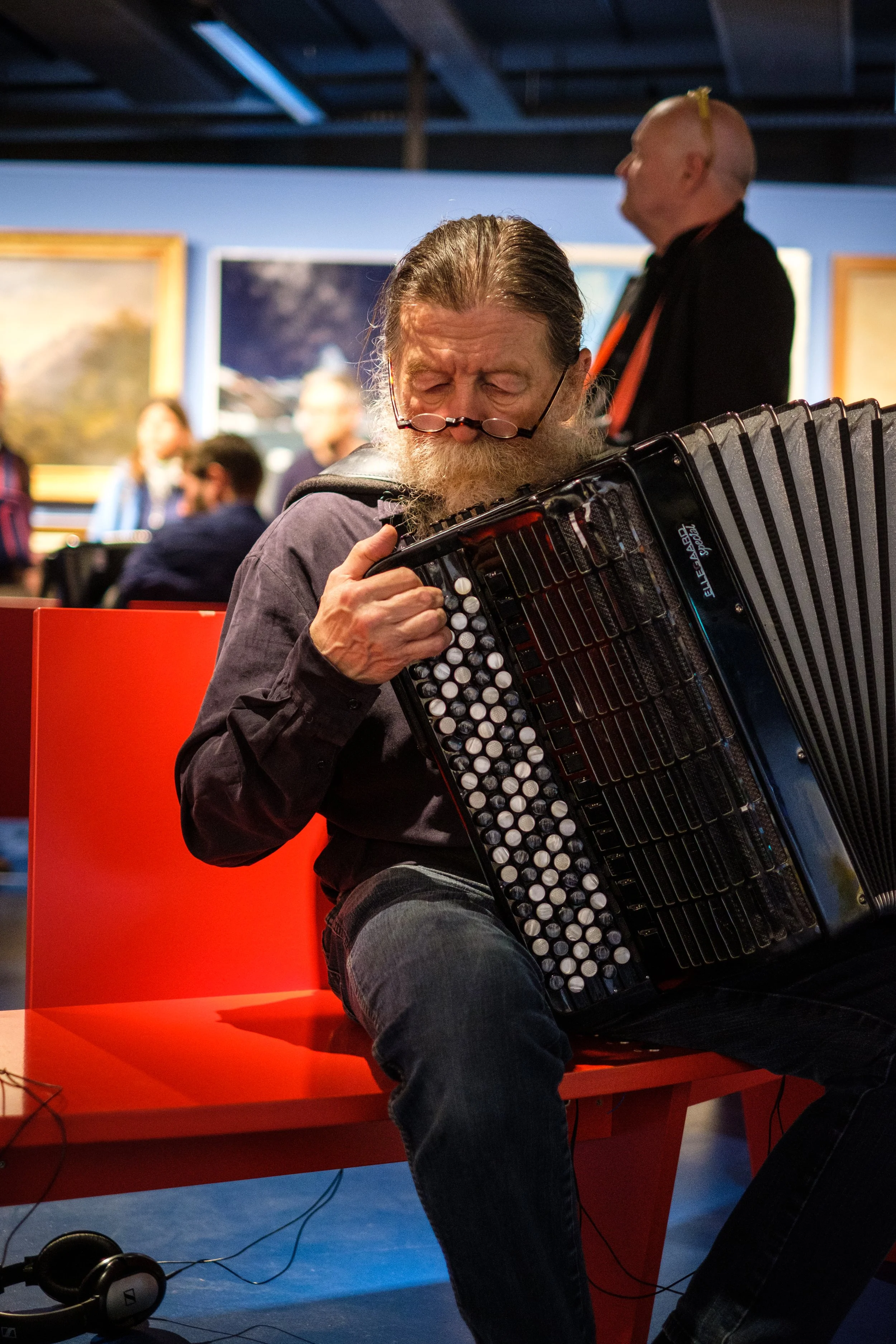 Ein älterer Mann mit Brille spielt auf einem Akkordeon in einem Kunstmuseum, während im Hintergrund andere Besucher die ausgestellten Gemälde betrachten.