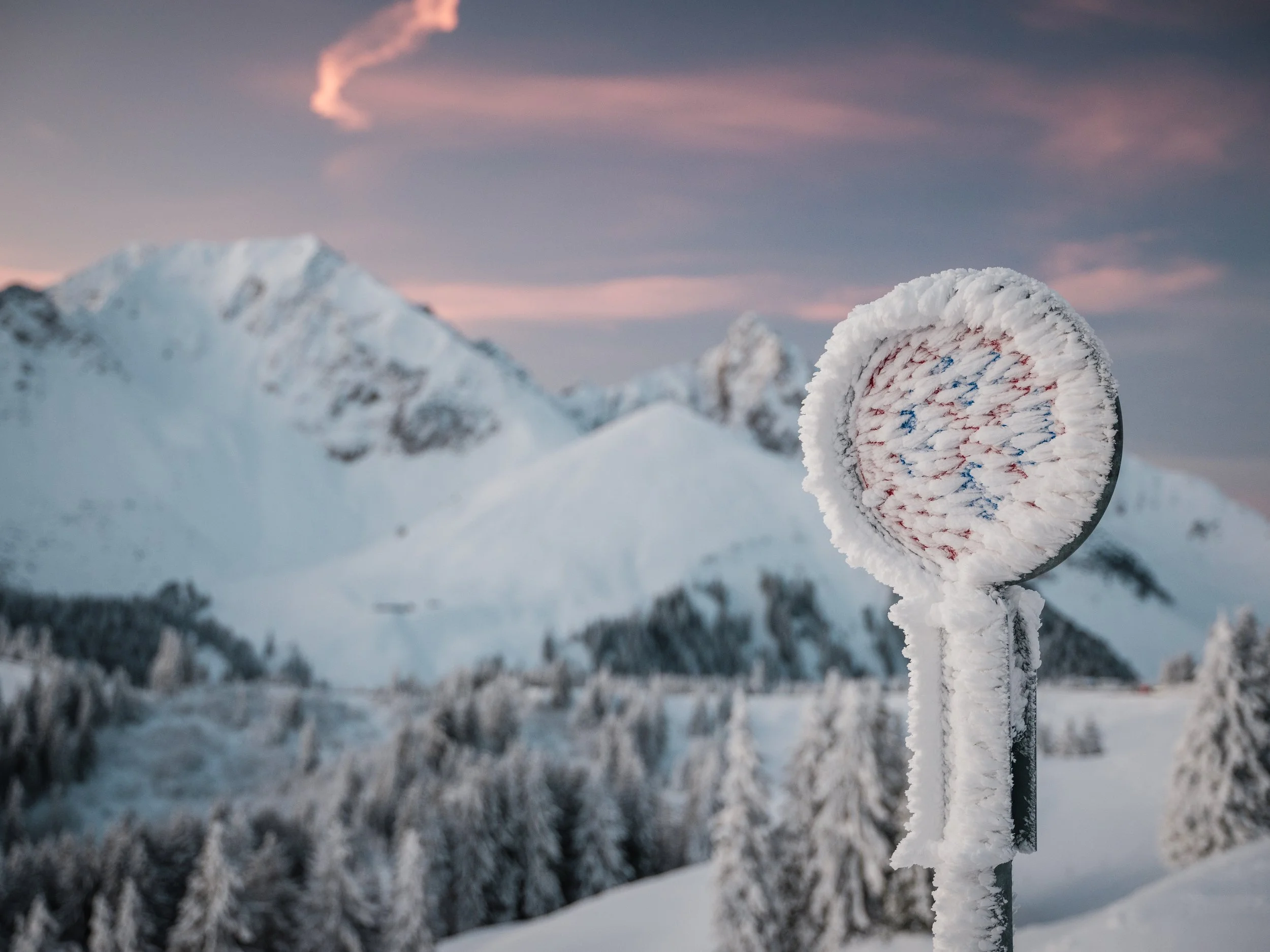 Schneebedeckter Bahnsteig mit gefrorenem Fahrkartenautomaten vor schneebedeckten Bergen bei Sonnenuntergang in den Alpen.