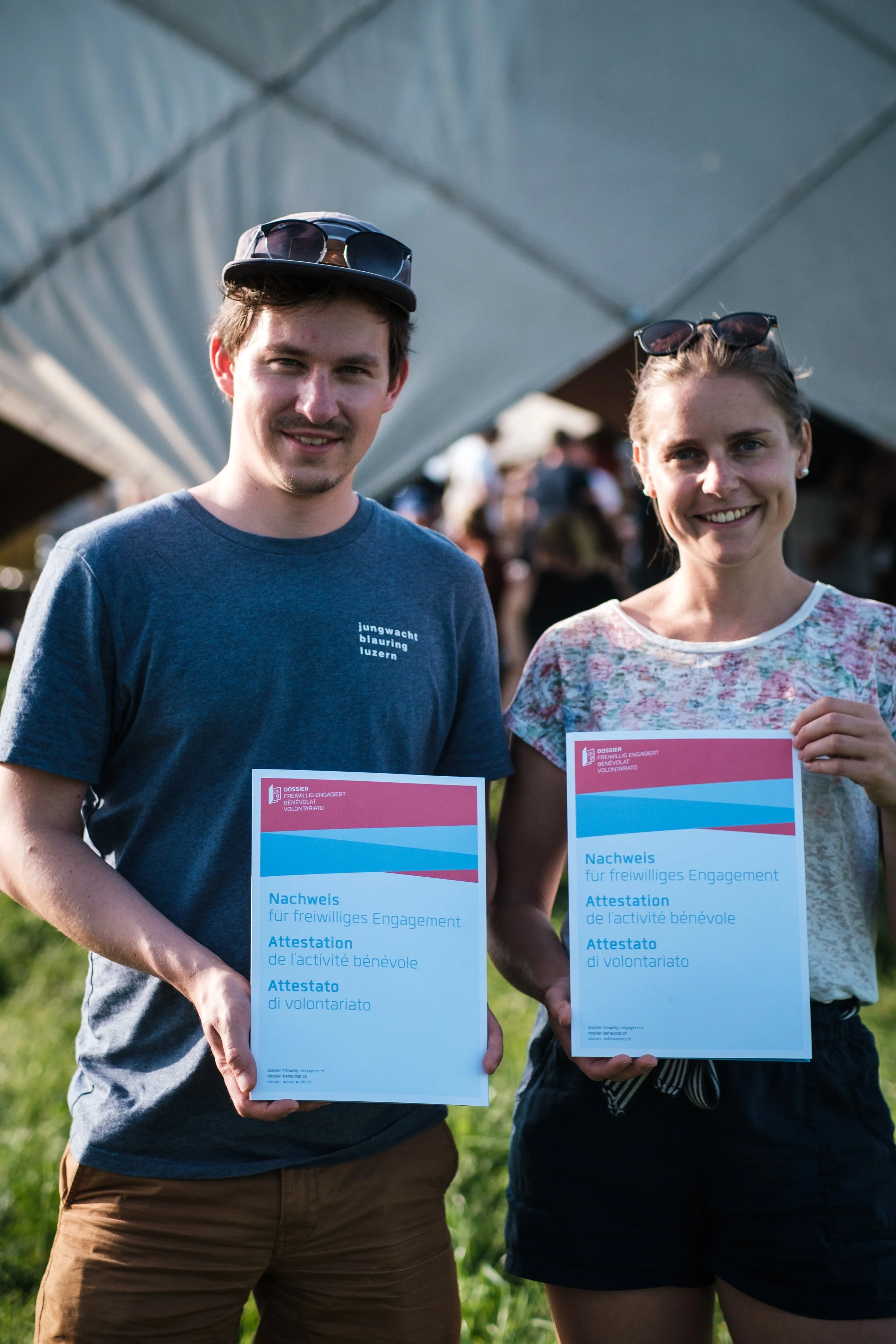 Zwei junge Menschen halten Zertifikate für freiwilliges Engagement bei einer Veranstaltung im Freien. Im Hintergrund sind mehrere Personen und ein Zelt sichtbar.
