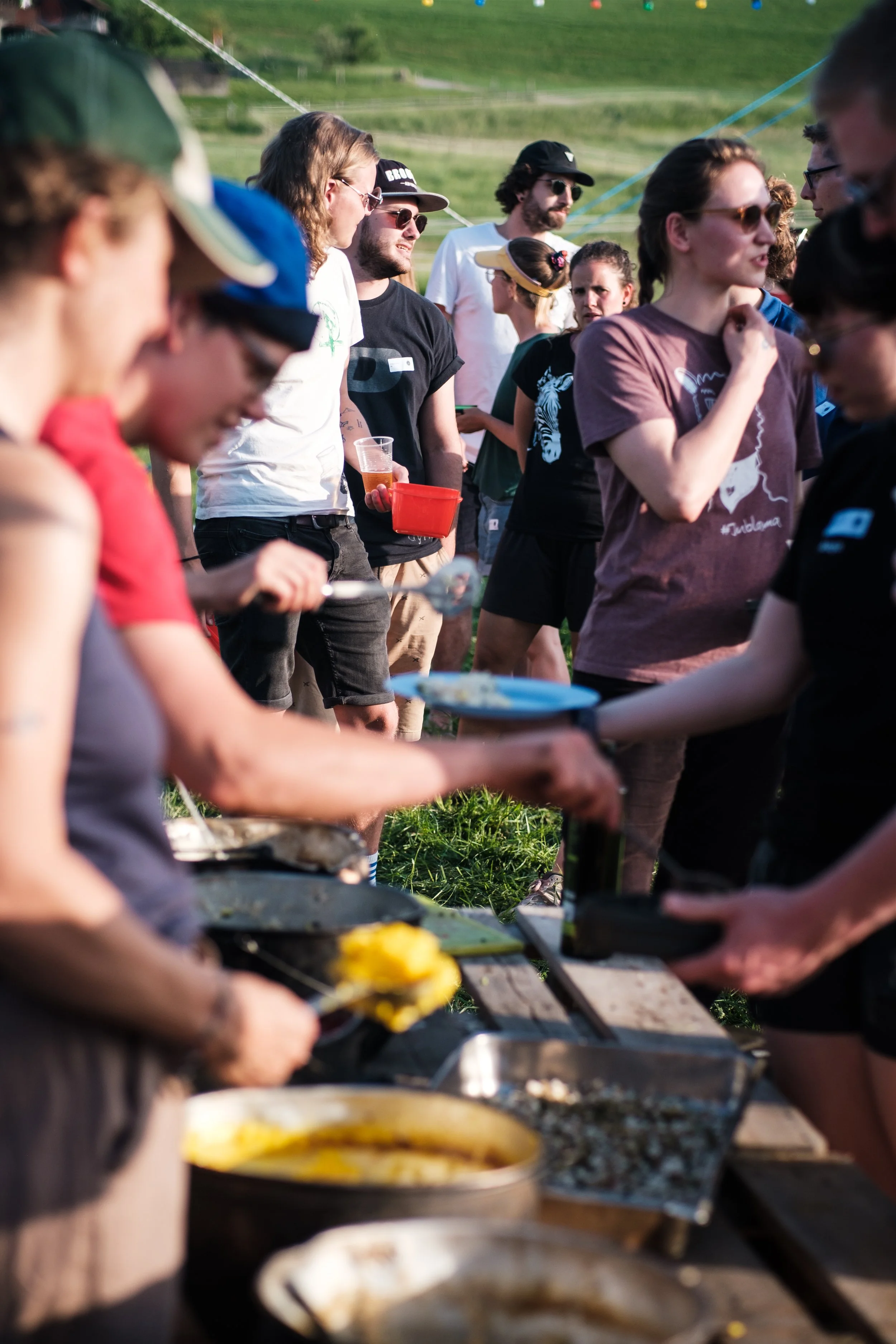 Menschen stehen in einer langen Reihe bei einem Buffet im Freien, wo Essen serviert wird, auf einer Wiese mit grünen Hügeln, während sie auf ihr Essen warten.
