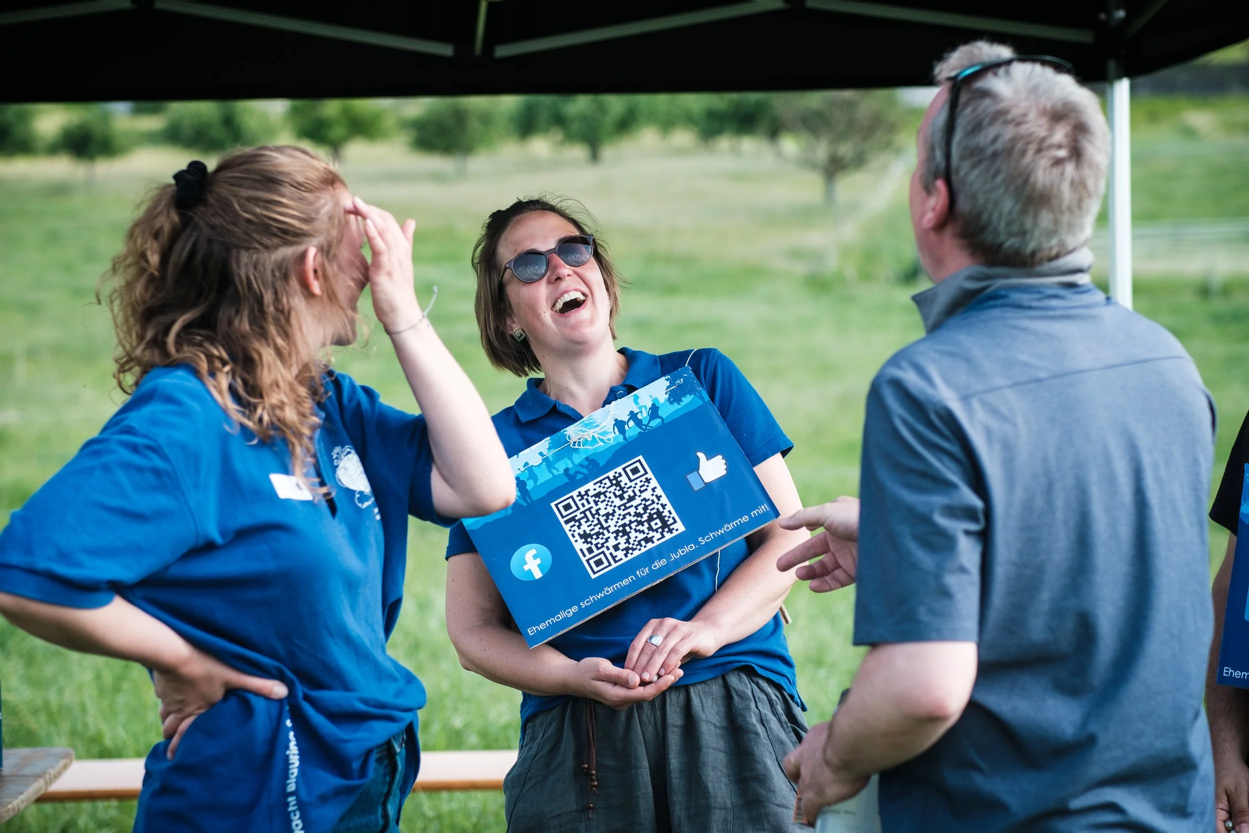Drei Menschen im Gespräch bei einem Outdoor-Event, eine Frau zeigt lachend auf eine QR-Code-Schild, während eine Frau mit Sonnenbrille und ein Mann mit Brille an der Kommunikation beteiligt sind.
