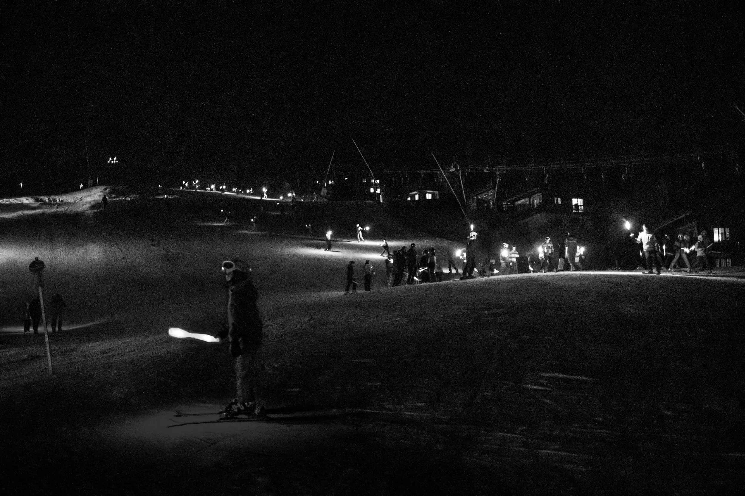 Skiort bei Nacht mit mehreren Skifahrern, die im Schneeland unterwegs sind, und Skilampen, die die Pisten beleuchten.