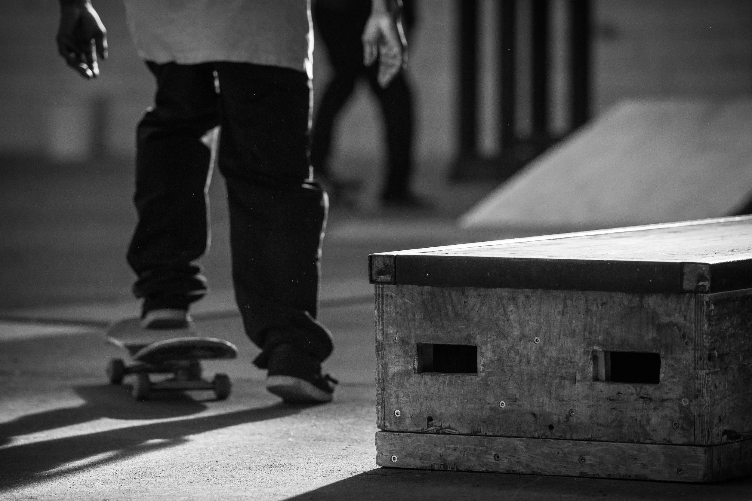Ein Junge auf einem Skateboard bei Nacht in einem Skatpark, im Vordergrund eine große Holzbox, im Hintergrund andere Skater, schwarz-weiße Aufnahme.